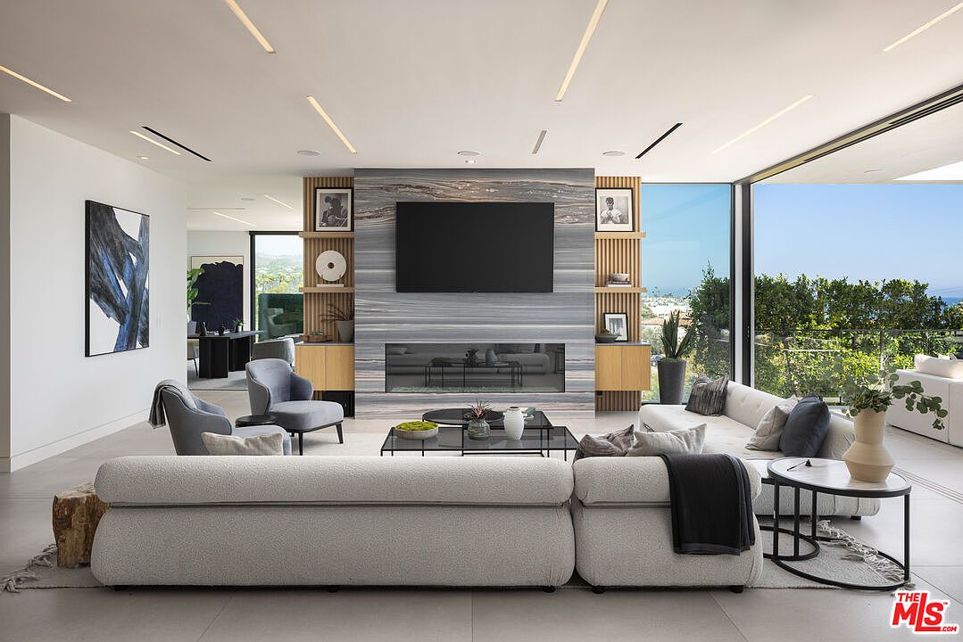 This sophisticated, open-concept living room features a striking floor-to-ceiling stone fireplace as its centerpiece, flanked by custom wood shelving. A large, plush sectional sofa and modern armchairs create a comfortable seating area, while expansive floor-to-ceiling glass walls offer a seamless transition to the outdoor scenery. The space is defined by its clean, contemporary aesthetic, recessed linear lighting, and a neutral color palette that emphasizes luxury and relaxation.