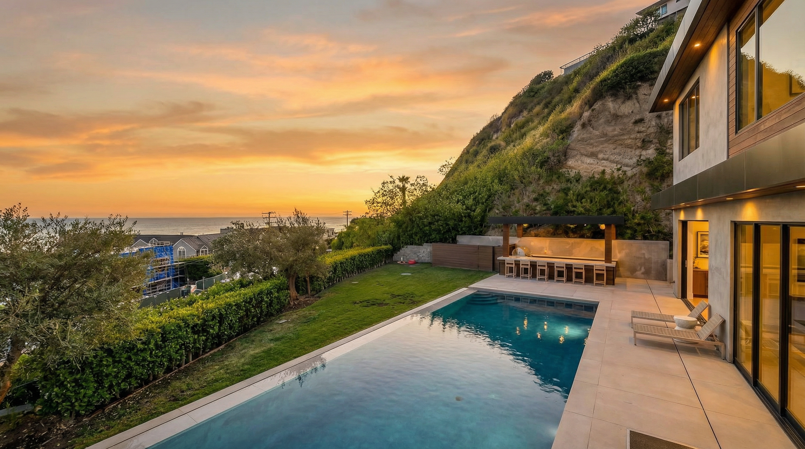 This elevated perspective captures a luxurious backyard oasis featuring a sleek, rectangular infinity-edge swimming pool as the centerpiece. The space includes a manicured lawn, a covered outdoor kitchen with bar seating, and a paved lounge area, all set against a backdrop of coastal cliffs and the Pacific Ocean. The scene exudes a high-end, serene atmosphere perfect for outdoor entertaining and relaxation.