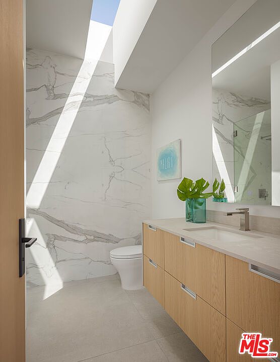 This modern, minimalist bathroom features a striking floor-to-ceiling marble accent wall illuminated by a dramatic skylight. The space includes a sleek, light-wood vanity with a clean countertop, a contemporary faucet, and a large mirror that reflects the bright, airy atmosphere. The overall aesthetic is sophisticated and serene, emphasizing high-end materials and natural light.