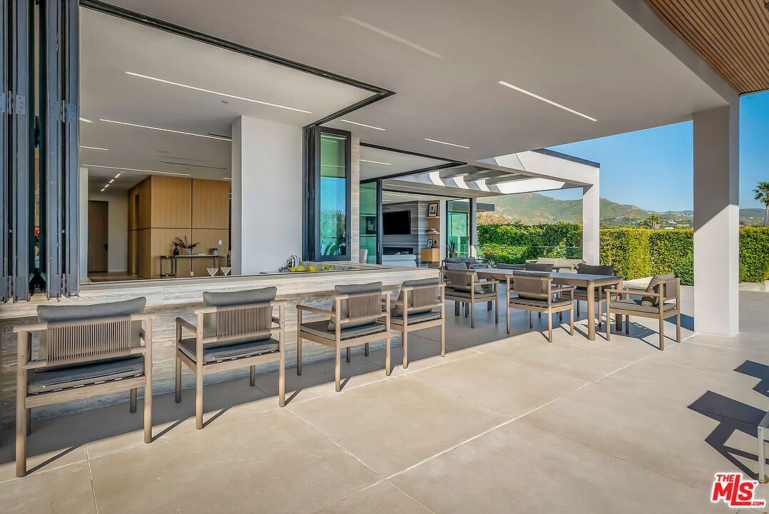 This expansive, modern covered patio features a seamless indoor-outdoor transition with large folding glass doors that open into the home's interior. The space is furnished with a sleek stone-topped bar with four chairs and a separate dining area, all set against a backdrop of lush green hedges and distant mountain views. The clean lines, neutral color palette, and recessed lighting create a sophisticated and inviting atmosphere perfect for luxury outdoor entertaining.
