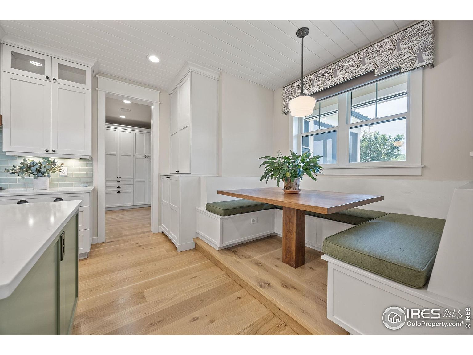 This is a bright and inviting dining nook featuring a built-in bench seating area with green cushions and a wooden table. A large window provides natural light, complemented by a stylish pendant light fixture. The space is adjacent to the kitchen and showcases a clean, modern design with white cabinetry and light wood flooring.