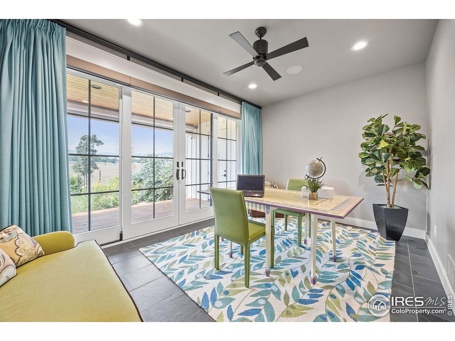 This is an interior shot of a home office featuring a desk with a laptop, two chairs, and a globe. Large sliding glass doors provide ample natural light and a view of the outdoors. The room is decorated with a colorful rug and a potted plant, creating a bright and inviting workspace.