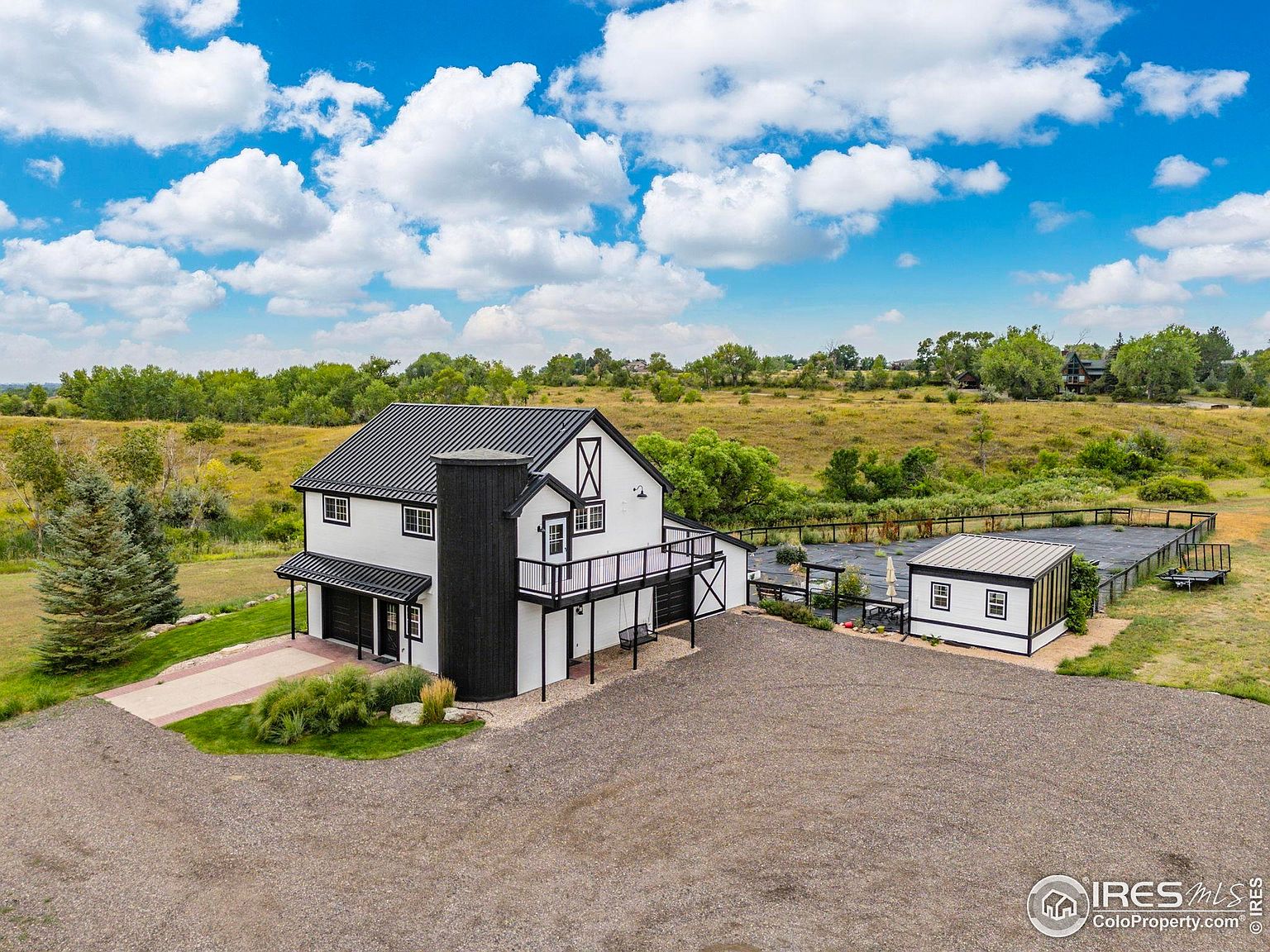 This aerial shot showcases a modern farmhouse-style home with a black metal roof and white siding accented by black trim. The property features a large gravel driveway, a well-maintained yard, and a detached structure that could serve as a guest house or studio. The surrounding landscape includes open fields and mature trees, providing a sense of privacy and space.