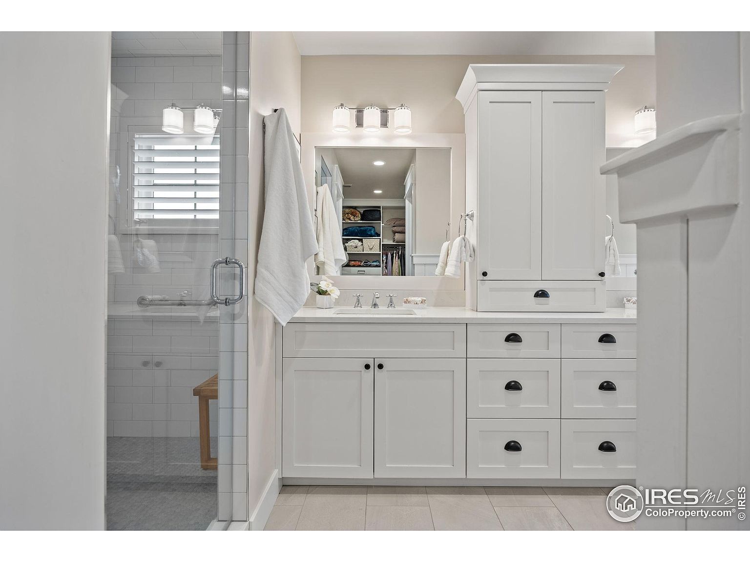 This is a well-lit primary bathroom featuring a white vanity with ample storage, including drawers and cabinets. A large mirror reflects the space, and modern lighting fixtures illuminate the countertop. The shower is visible through a glass door, adding to the bright and clean aesthetic of the bathroom.