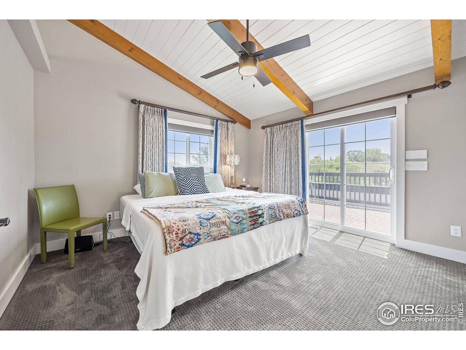 This is a primary bedroom featuring a bed with a white comforter and decorative throw, flanked by windows with curtains. The room has a vaulted ceiling with exposed wooden beams and a ceiling fan. A sliding glass door leads to a balcony, and the room is carpeted in a neutral tone, creating a cozy and inviting atmosphere.