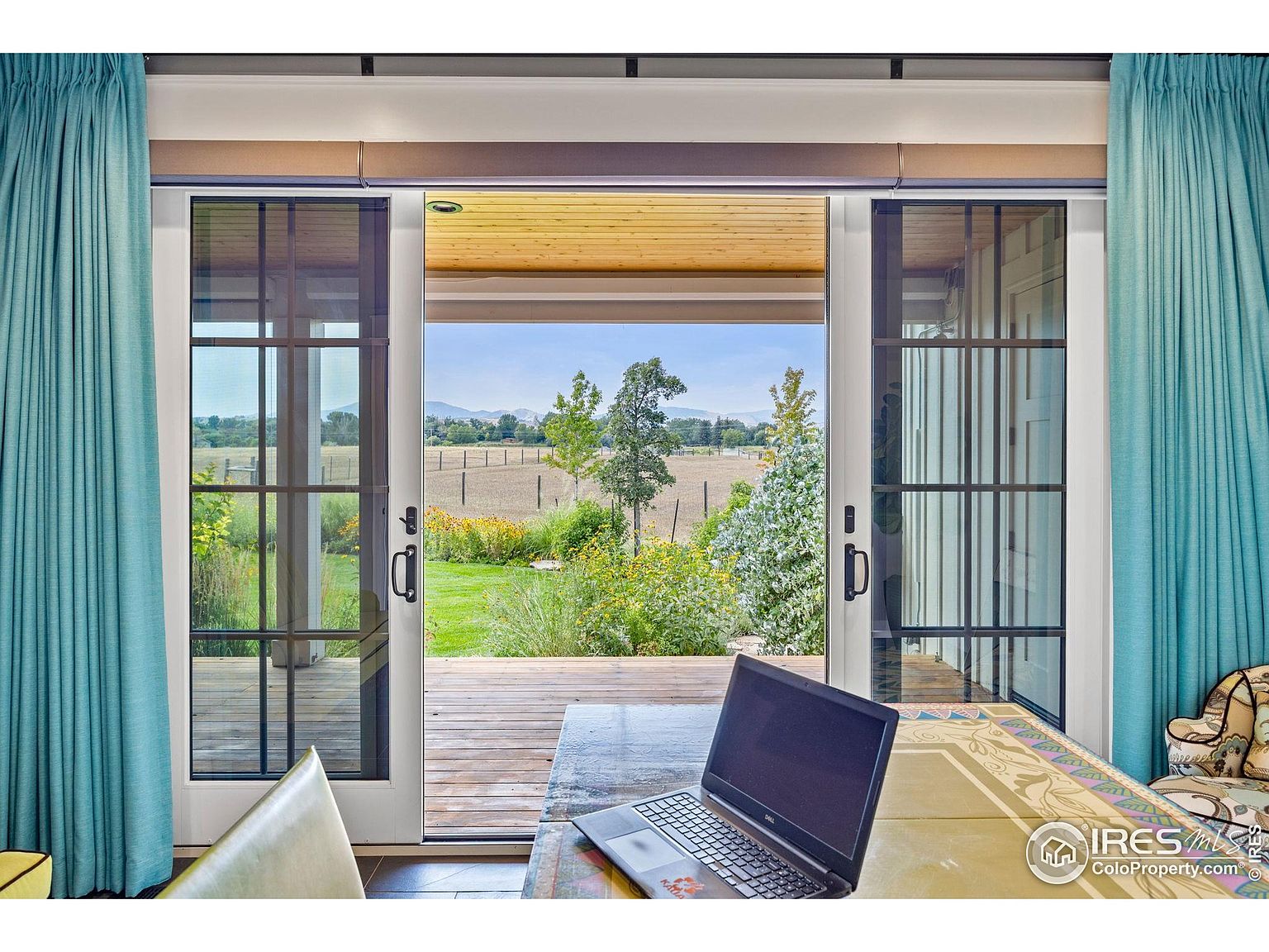 The image showcases an interior office space with a focus on the view through a large sliding glass door. The door opens onto a wooden deck and a lush garden, extending to a field and distant mountains. A laptop sits on a table in the foreground, suggesting a workspace with a connection to the outdoors, enhanced by the natural light and serene landscape.