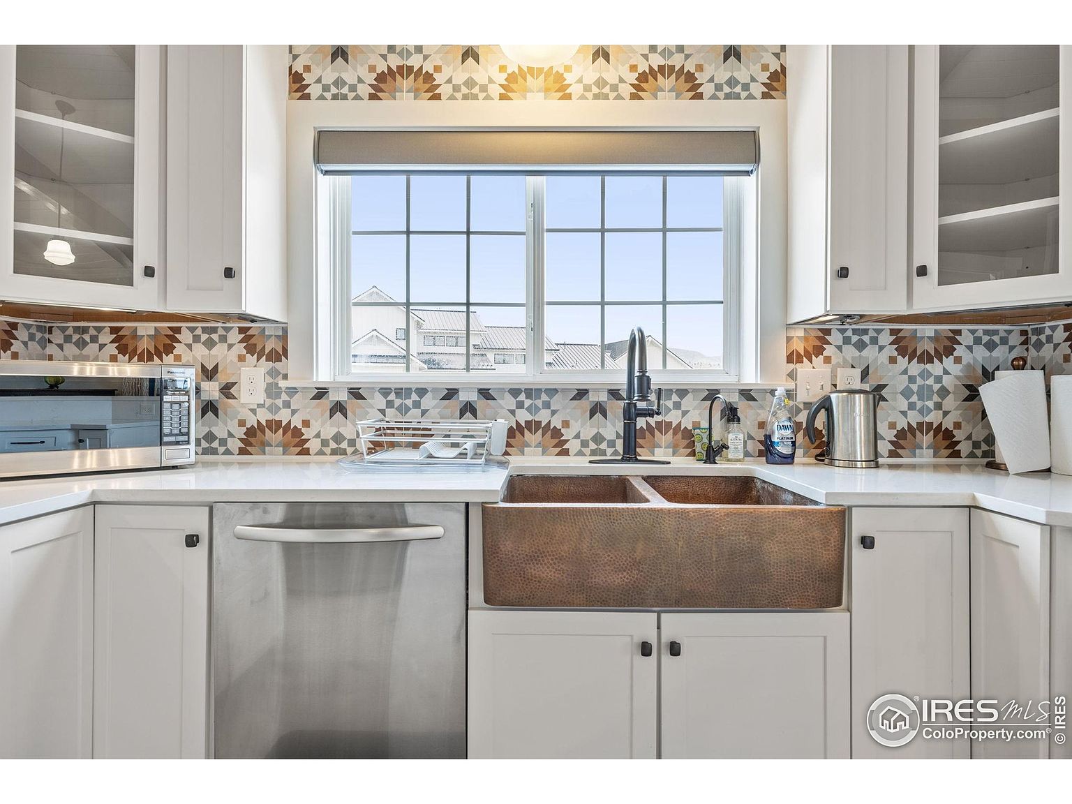 This is a well-lit kitchen featuring white cabinetry, stainless steel appliances, and a copper farmhouse sink. The backsplash is a patterned tile, and a large window provides natural light. The kitchen has a clean and modern aesthetic, with a focus on functionality and style.