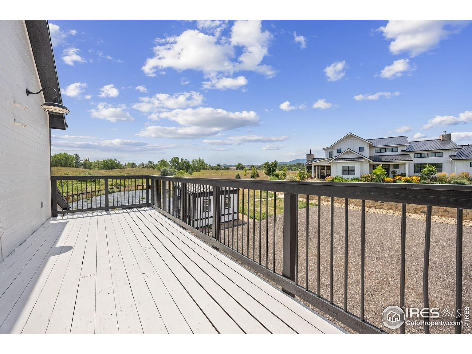 This image showcases a spacious deck with white painted wooden planks and a dark railing, offering a view of the property's exterior. A modern farmhouse-style house is visible in the background, complemented by a gravel driveway and landscaping. The scene is set under a bright, partly cloudy sky, creating an inviting outdoor living space.
