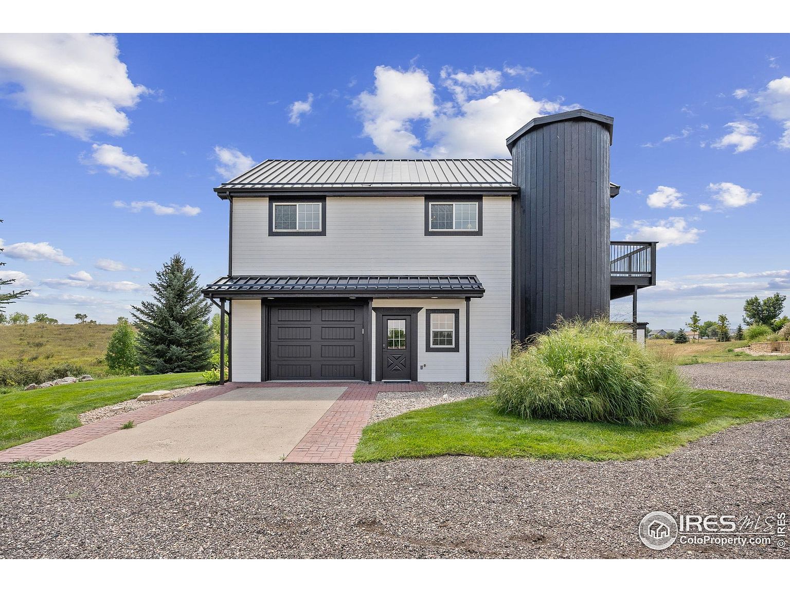 This is a front exterior view of a modern farmhouse-style home. The house features light gray siding with black trim, a metal roof, and a unique silo-like structure attached to the side with a balcony. The property includes a well-maintained lawn and a gravel driveway, creating a welcoming and stylish curb appeal.