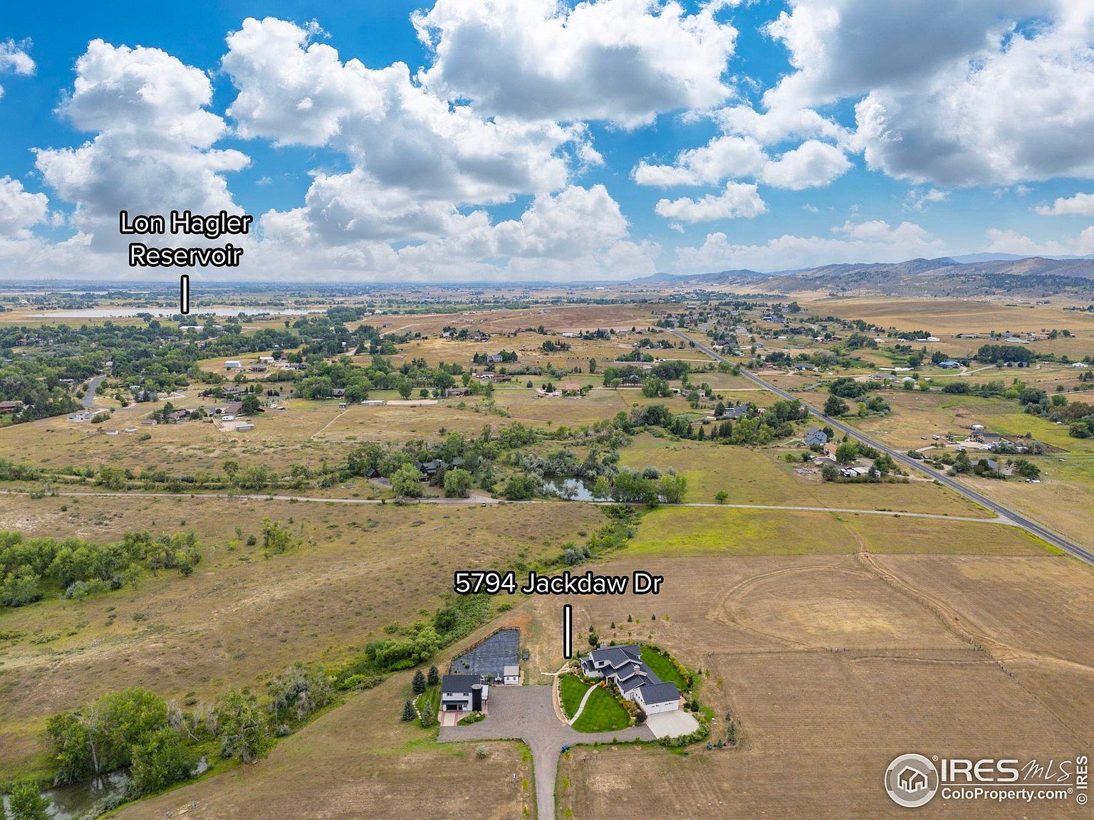 This aerial view showcases a property at 5794 Jackdaw Dr, featuring two houses with dark roofs and well-maintained yards. The landscape includes open fields and scattered trees, with a reservoir visible in the distance under a partly cloudy sky. The overall impression is one of spaciousness and rural tranquility.