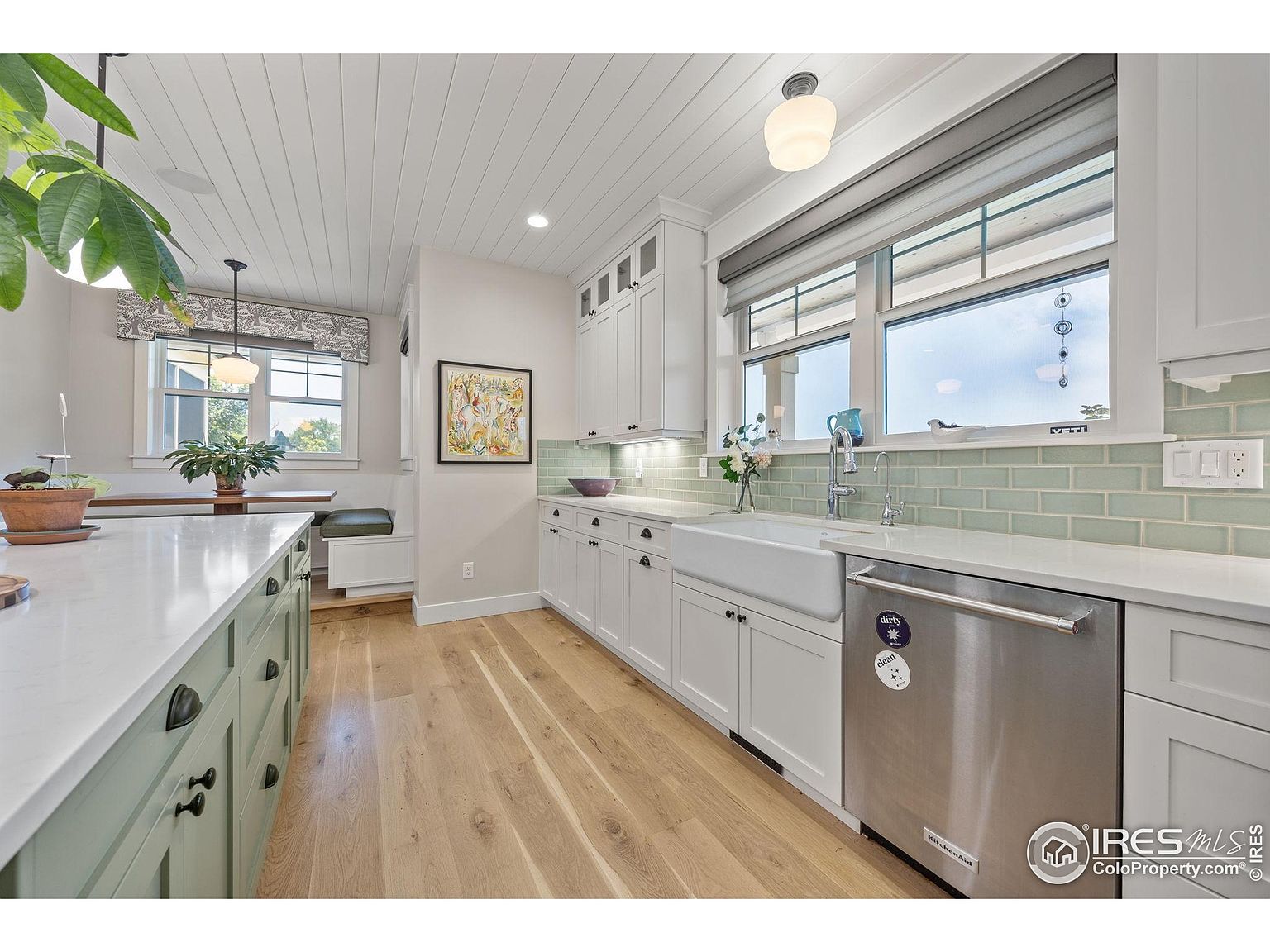 This is a bright and airy kitchen featuring white cabinetry, a farmhouse sink, and stainless steel appliances. The light wood flooring complements the white countertops and the light green backsplash, creating a fresh and inviting space. A large window provides natural light, and the overall design is both modern and charming.
