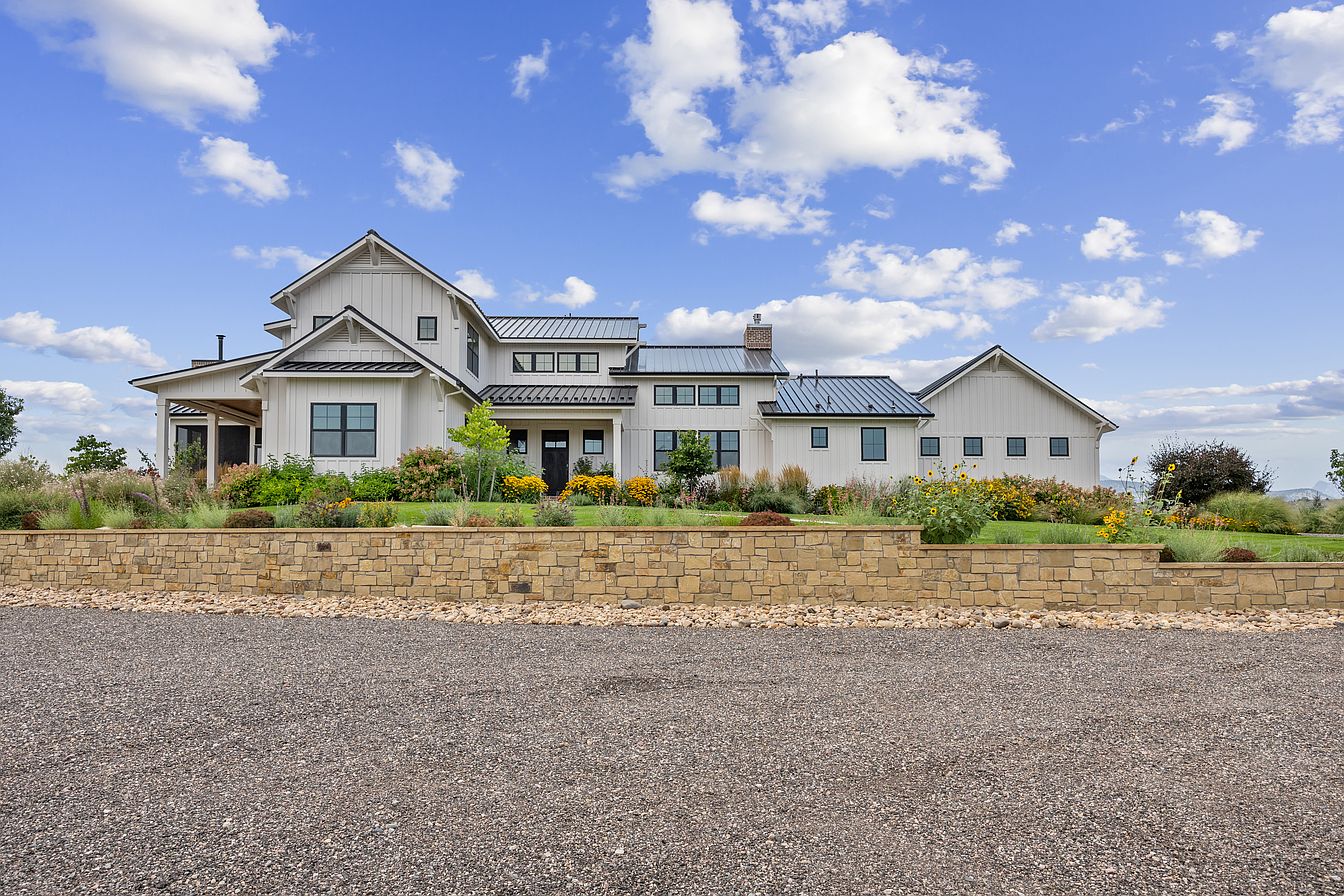 This image showcases the front exterior of a modern farmhouse-style home. The house features white siding, black framed windows, and a dark metal roof. A low stone retaining wall borders a well-maintained front yard with colorful flowers and mature landscaping, creating an inviting curb appeal.