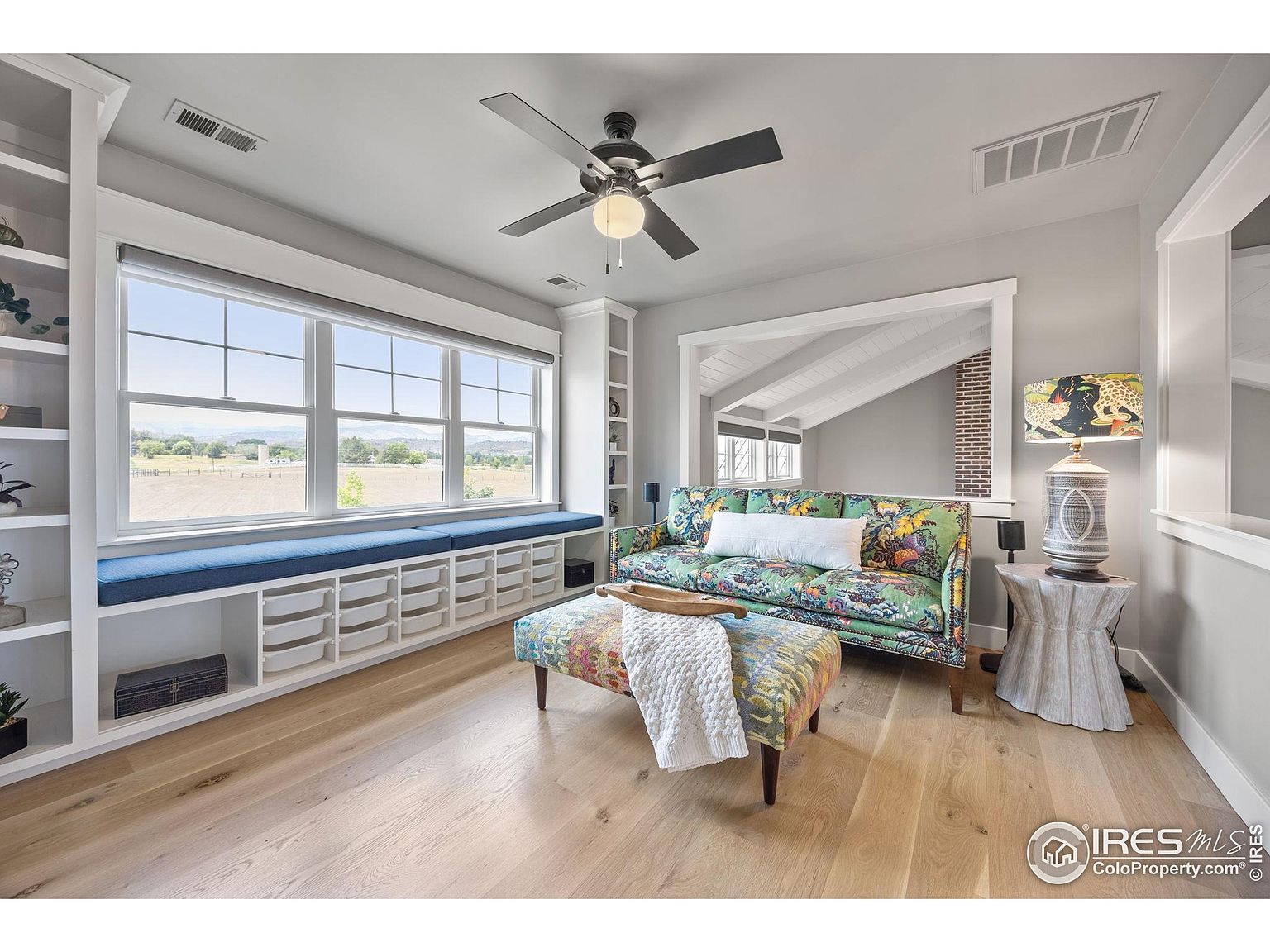This is an interior shot of a living room featuring a large window with a built-in bench and storage. The room is decorated with a colorful sofa and ottoman, complemented by a unique side table and lamp. The hardwood floors and neutral wall color create a bright and inviting space.