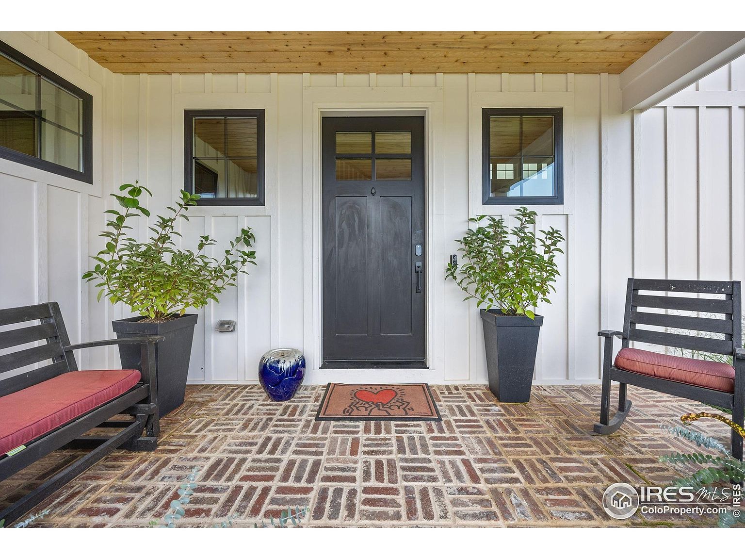 This is a welcoming entryway featuring a brick-paved porch with a heart-themed doormat leading to a dark-colored front door with glass panes. Flanking the door are matching black-framed windows and potted plants, adding symmetry and curb appeal. Two black rocking chairs with red cushions sit on either side, creating an inviting space.