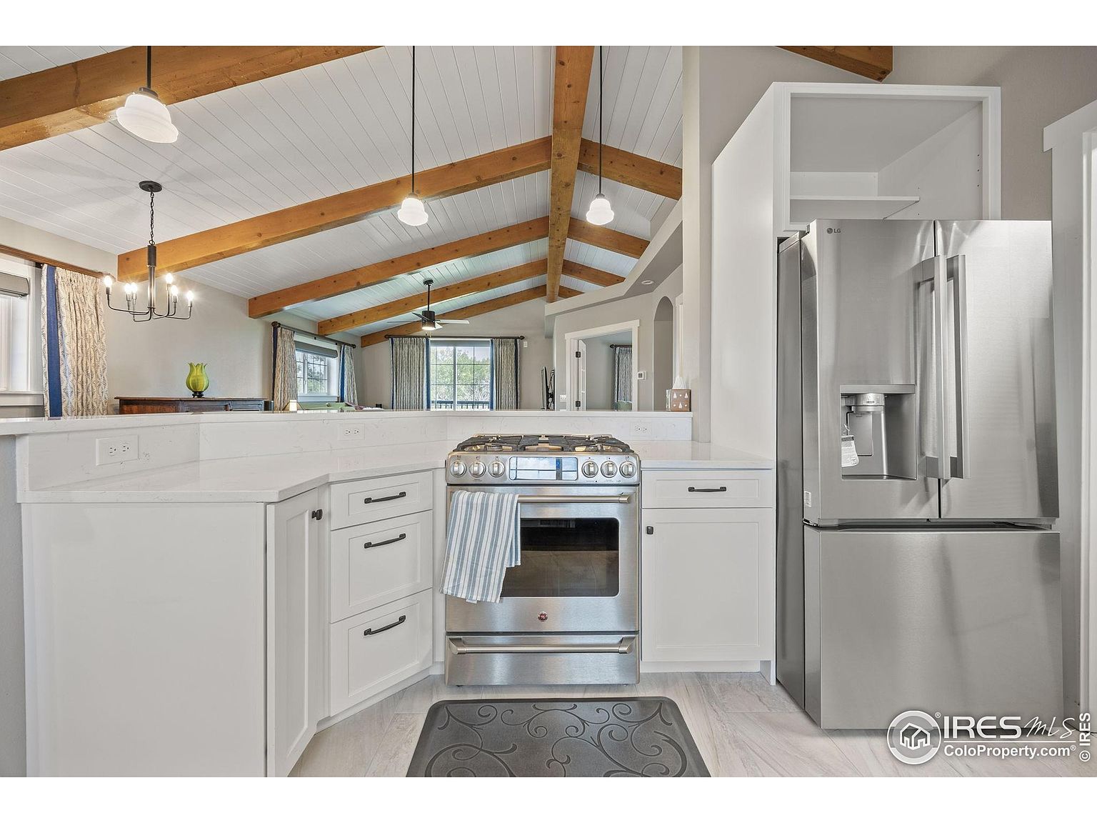 This is a bright kitchen featuring white cabinets, stainless steel appliances, and a vaulted ceiling with exposed wooden beams. The kitchen includes a stainless steel range and refrigerator, with a view into an adjacent living area. The overall impression is clean and modern with rustic touches.
