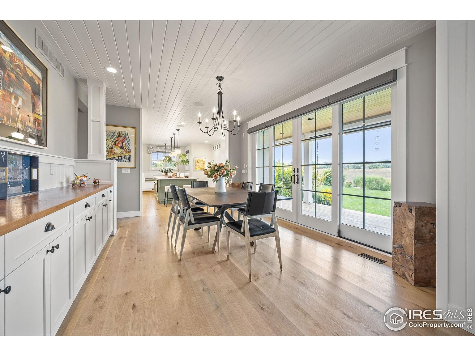 This is an interior shot of a dining room featuring a wooden dining table with black chairs, a chandelier, and large sliding glass doors that lead to an outdoor area. The room has light wood flooring and white paneled ceilings, creating a bright and airy atmosphere. The space is well-lit and appears to be part of an open-concept design, possibly connected to the kitchen.