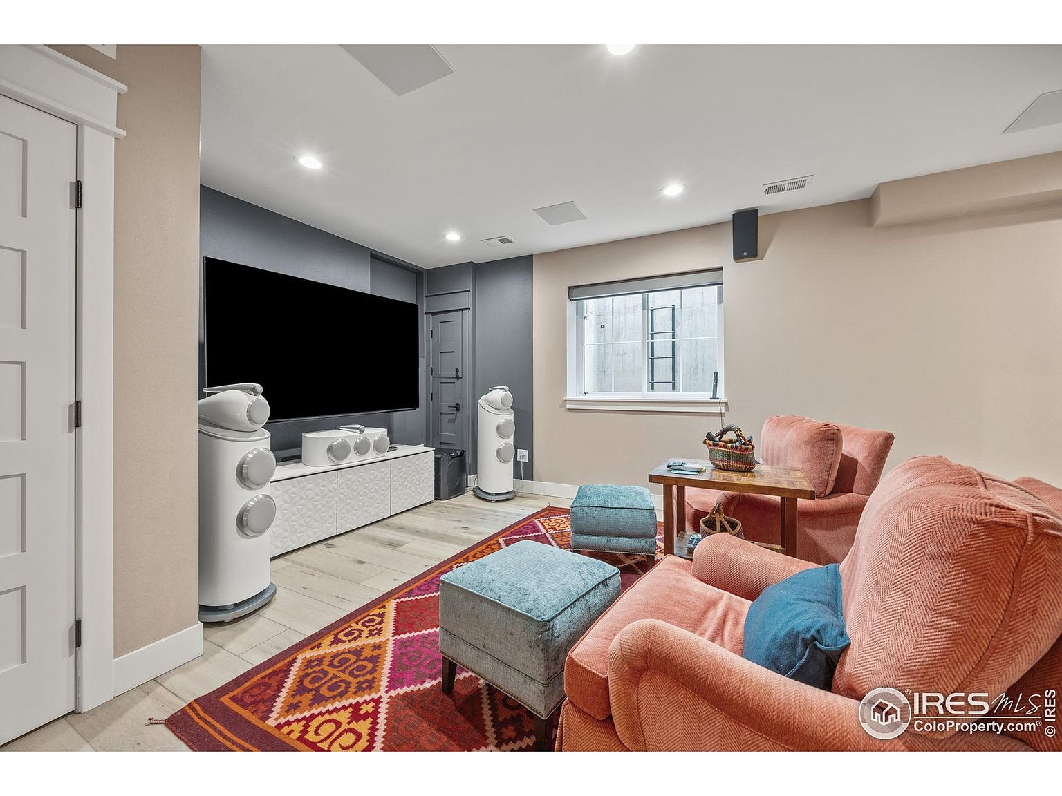 This is an interior shot of a living room featuring a large television mounted on a gray accent wall, flanked by modern white speakers. The room is furnished with comfortable-looking armchairs, blue ottomans, and a patterned rug, creating a cozy and inviting atmosphere. A window provides natural light, and the overall design suggests a modern and comfortable entertainment space.