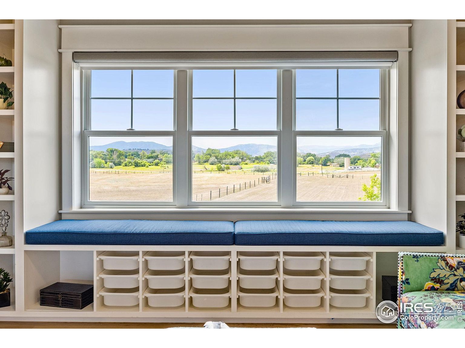 This interior shot showcases a charming sunroom or conservatory area featuring a large window seat with a blue cushion. Below the window seat, there are built-in storage compartments filled with white bins, offering practical organization. The window provides a scenic view of a field and distant mountains, enhancing the room's appeal and creating a serene atmosphere.