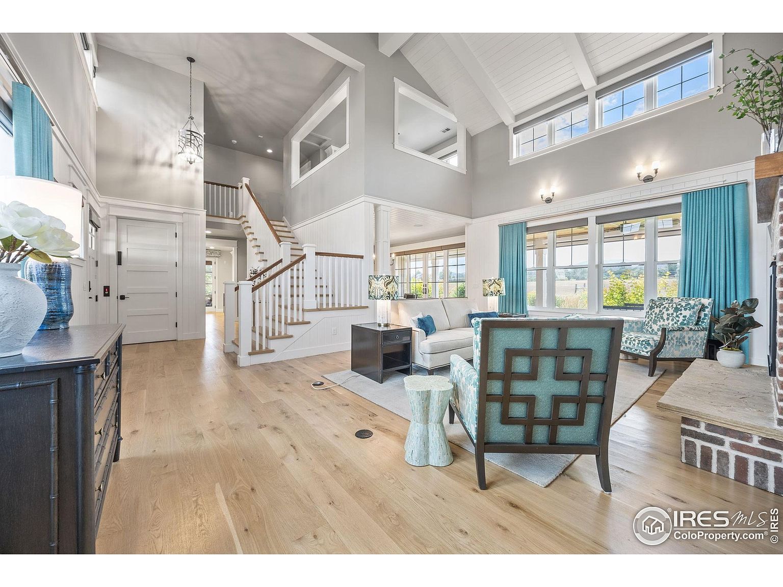 This is an interior shot of a living room featuring a high vaulted ceiling with exposed beams and large windows that provide ample natural light. The room is decorated in a modern style with a neutral color palette accented by blue curtains and patterned chairs. A staircase is visible in the background, adding architectural interest to the space.
