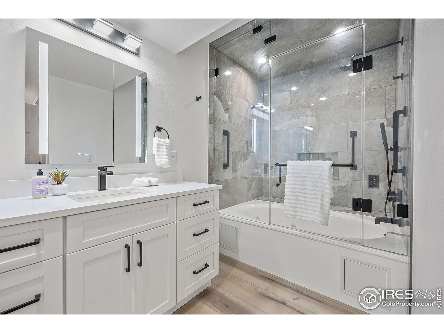 This is a well-lit primary bathroom featuring a modern design with white cabinetry and black hardware. A large, illuminated mirror hangs above the sink, and a glass-enclosed bathtub and shower combination is visible in the background. The bathroom has a clean and luxurious feel, enhanced by the light wood flooring.