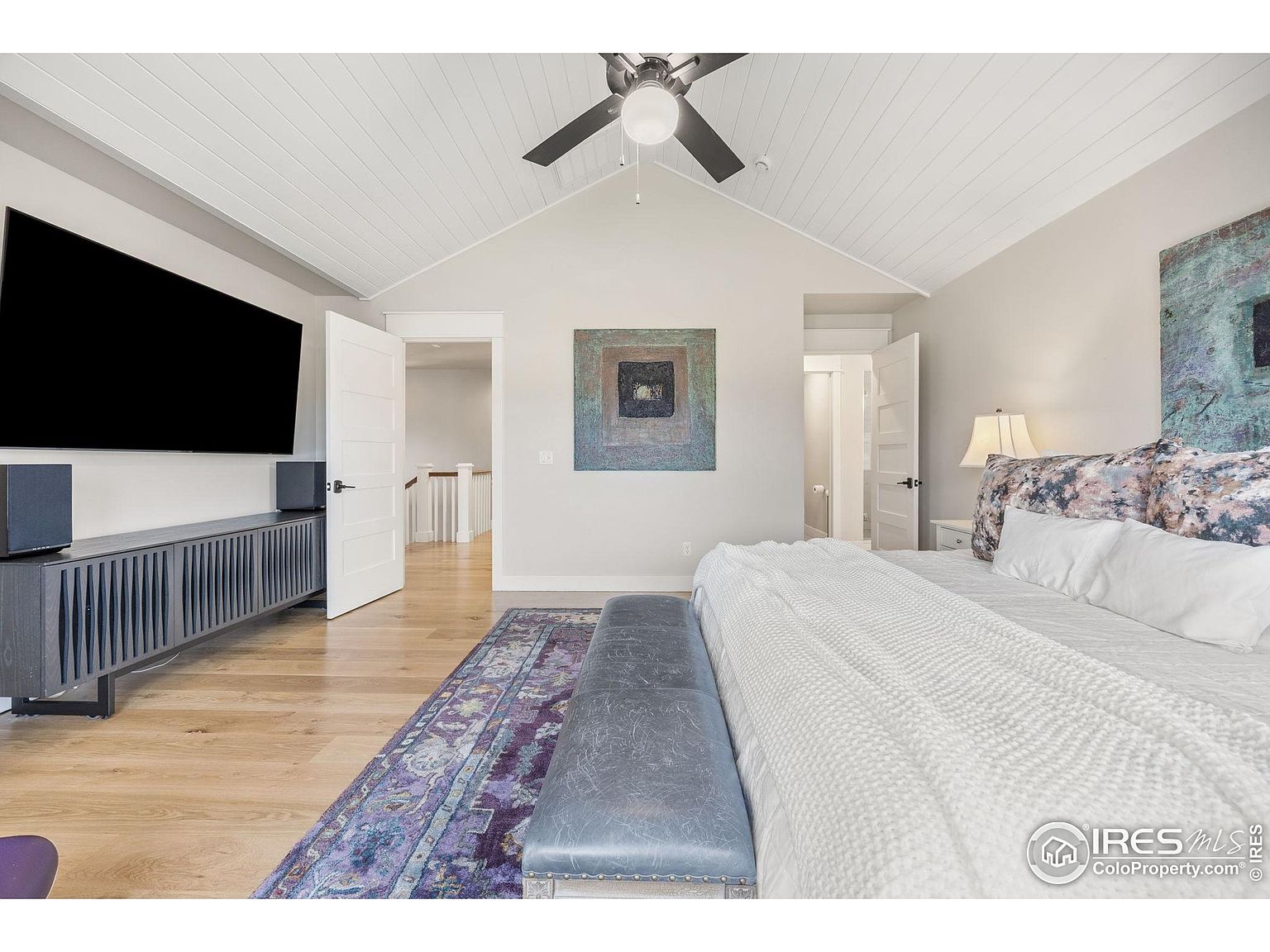 This is a primary bedroom featuring a vaulted ceiling with white wood paneling and a ceiling fan. The room is decorated with neutral tones, a large bed with white bedding, and artwork on the walls. A console table with a large television sits against one wall, and doorways lead to other areas of the house, creating an open and airy feel.
