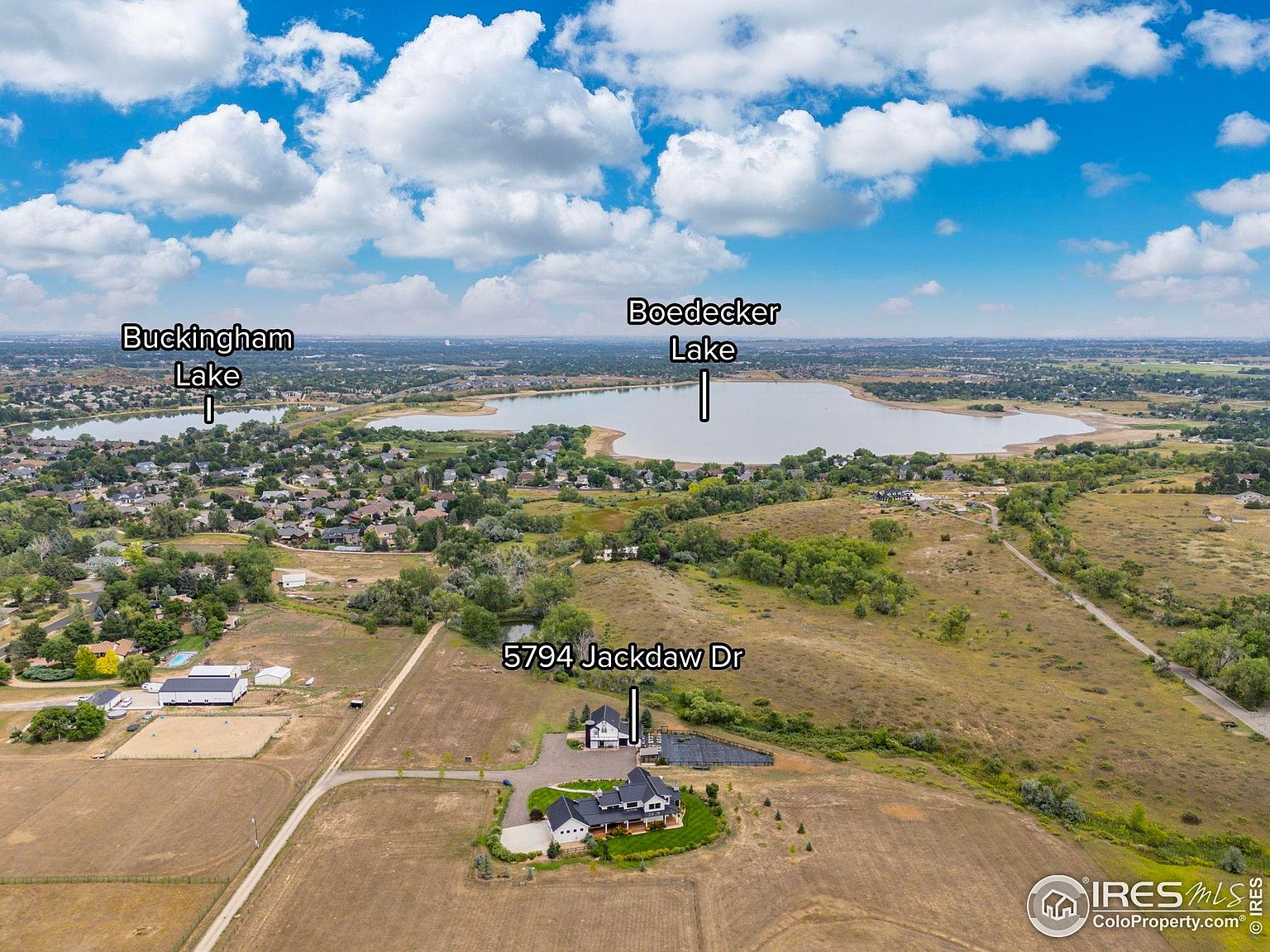 This aerial view showcases a sprawling property featuring a large, modern home with a dark roof and light-colored exterior, complemented by a smaller outbuilding. The residence is situated on a vast, mostly dry landscape with a long driveway leading to the house. In the distance, two lakes, Buckingham Lake and Boedecker Lake, add a scenic backdrop to the property, enhancing its appeal.