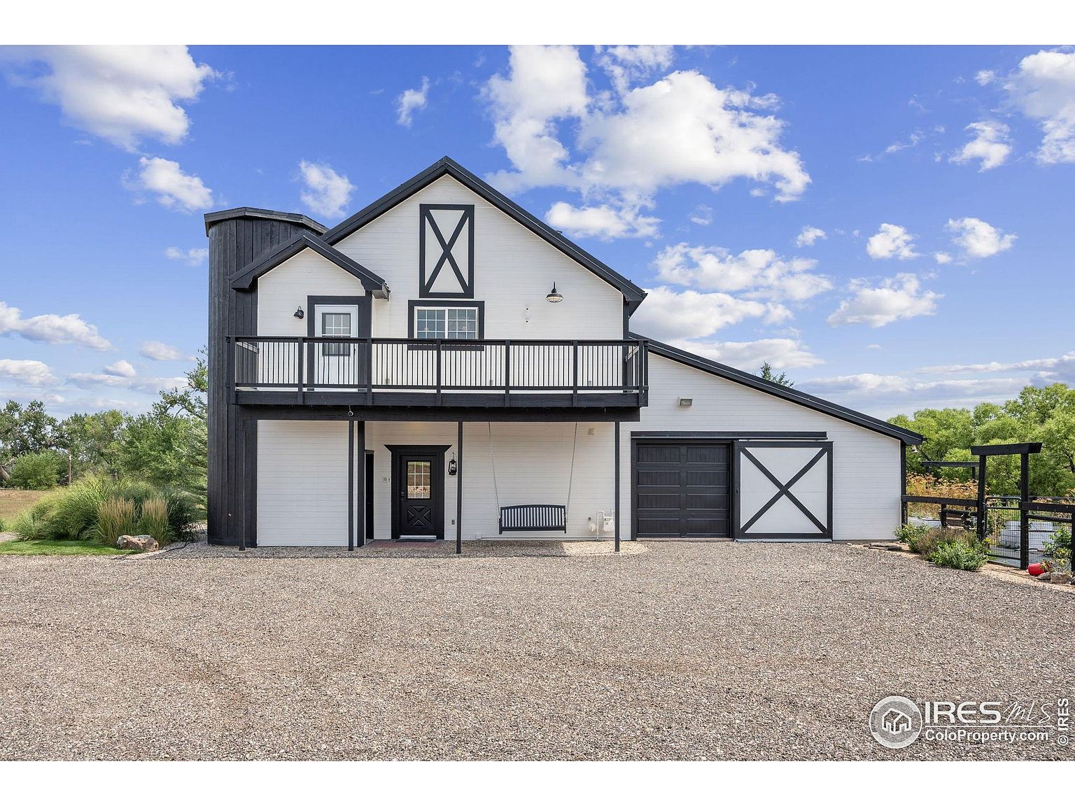 This is a front exterior view of a modern farmhouse-style home. The house features white siding with black trim, a second-story balcony, and a large gravel driveway. A porch swing hangs beneath the balcony, adding a touch of charm, and a sliding barn door is visible on the right side of the house.