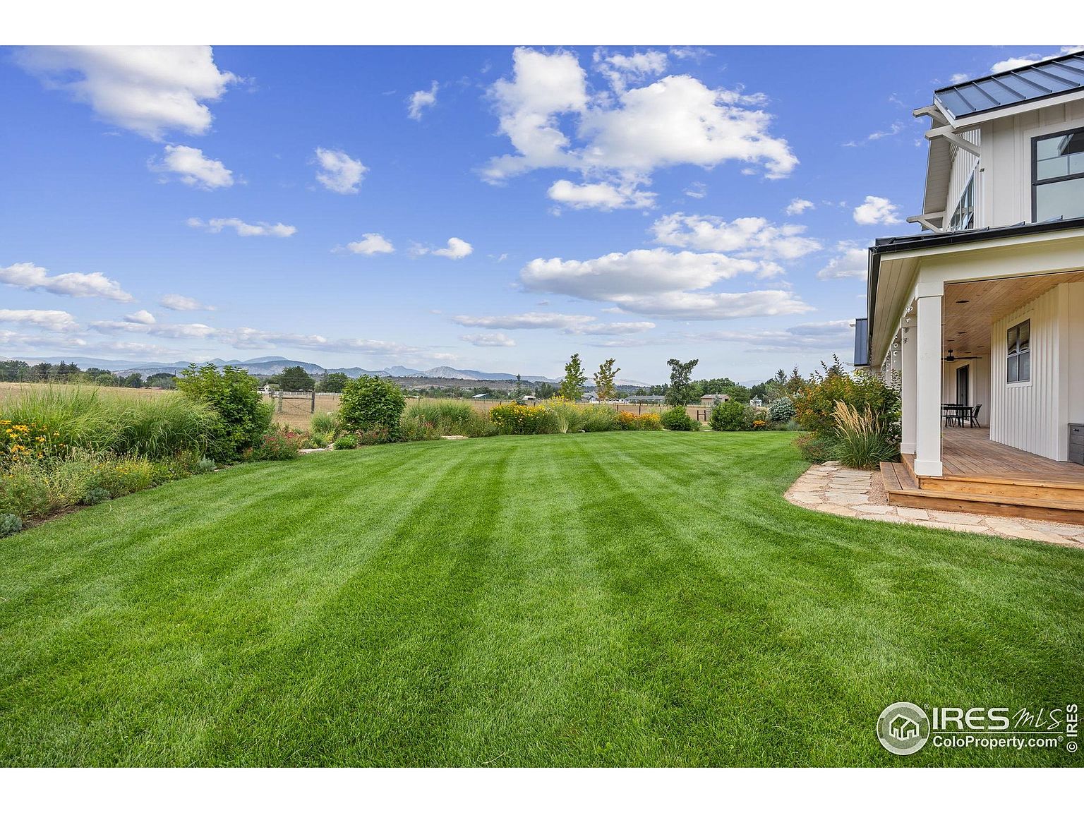 This image showcases a meticulously maintained backyard with a lush green lawn and well-manicured landscaping. The yard extends towards a distant view of fields and mountains under a partly cloudy sky. A portion of the house with a covered porch is visible on the right, suggesting a seamless transition between indoor and outdoor living.