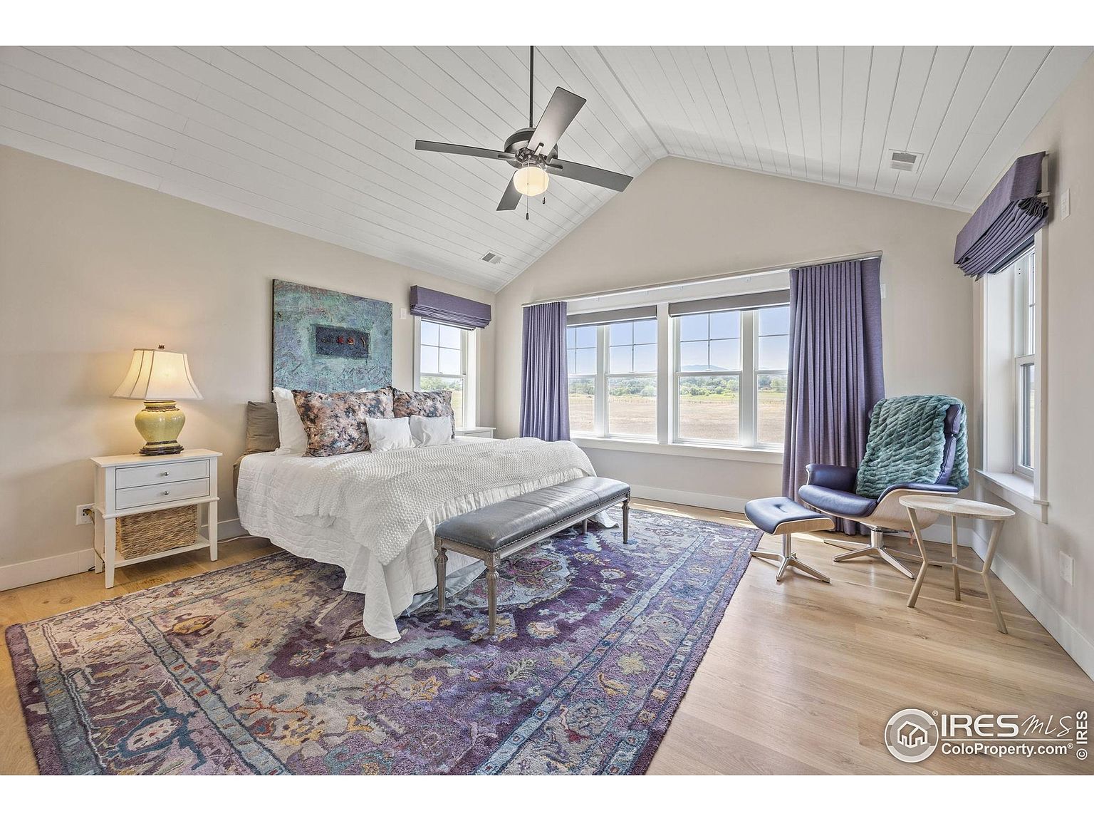 This is a primary bedroom featuring a king-size bed with a white comforter and decorative pillows, set against a backdrop of a blue abstract art piece. The room is well-lit by natural light streaming through multiple windows, which are adorned with purple curtains and shades. A patterned rug adds warmth and character to the light wood flooring, while a ceiling fan provides air circulation, creating a serene and inviting atmosphere.