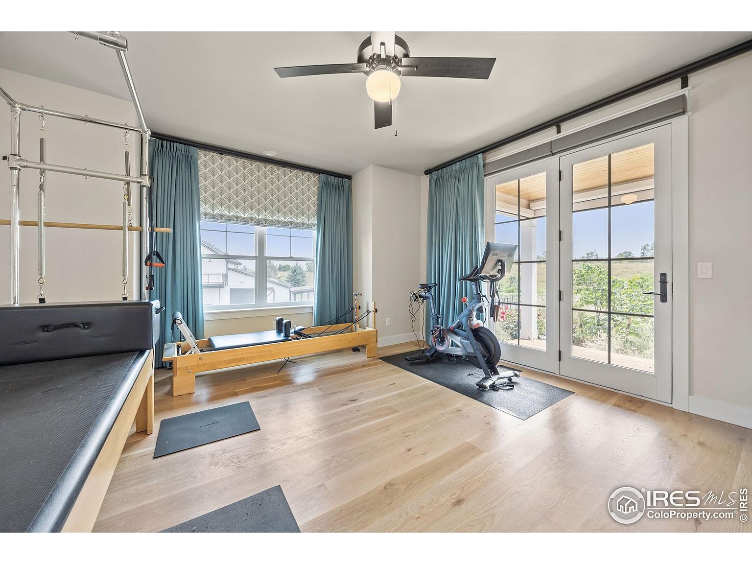 This is an interior shot of a home gym featuring a Pilates reformer, a stationary exercise bike, and yoga mats on a light hardwood floor. Natural light floods the room through a large window and French doors, which are adorned with blue curtains. A ceiling fan provides air circulation, creating a bright and inviting workout space.