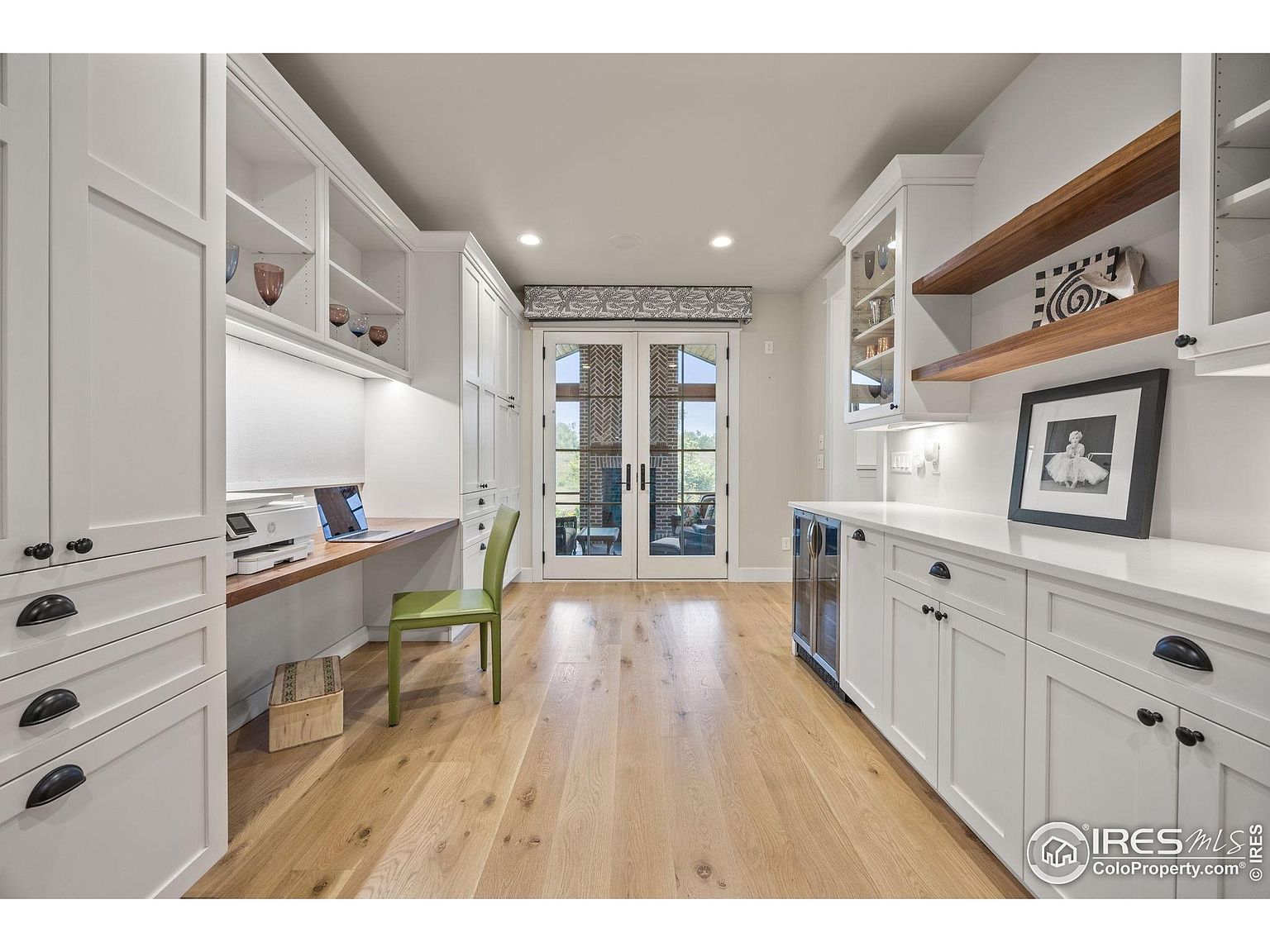 This is an interior shot of a well-organized home office or study area. The room features custom white cabinetry, a built-in desk with a wooden countertop, and a green chair. Double doors lead to an outdoor space, and the overall impression is one of functionality and style.