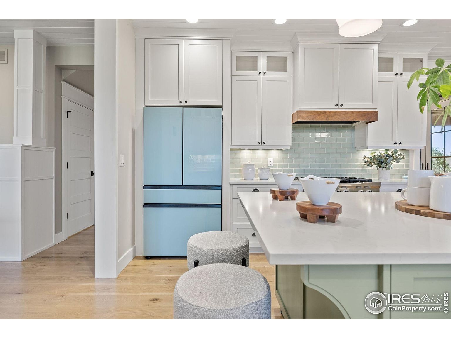 This is a bright and airy kitchen featuring white cabinetry, a light blue refrigerator, and a large island with a white countertop and sage green base. The backsplash is a light blue tile, and the overall style is modern and clean. The perspective is from the island, looking towards the refrigerator and cabinets.