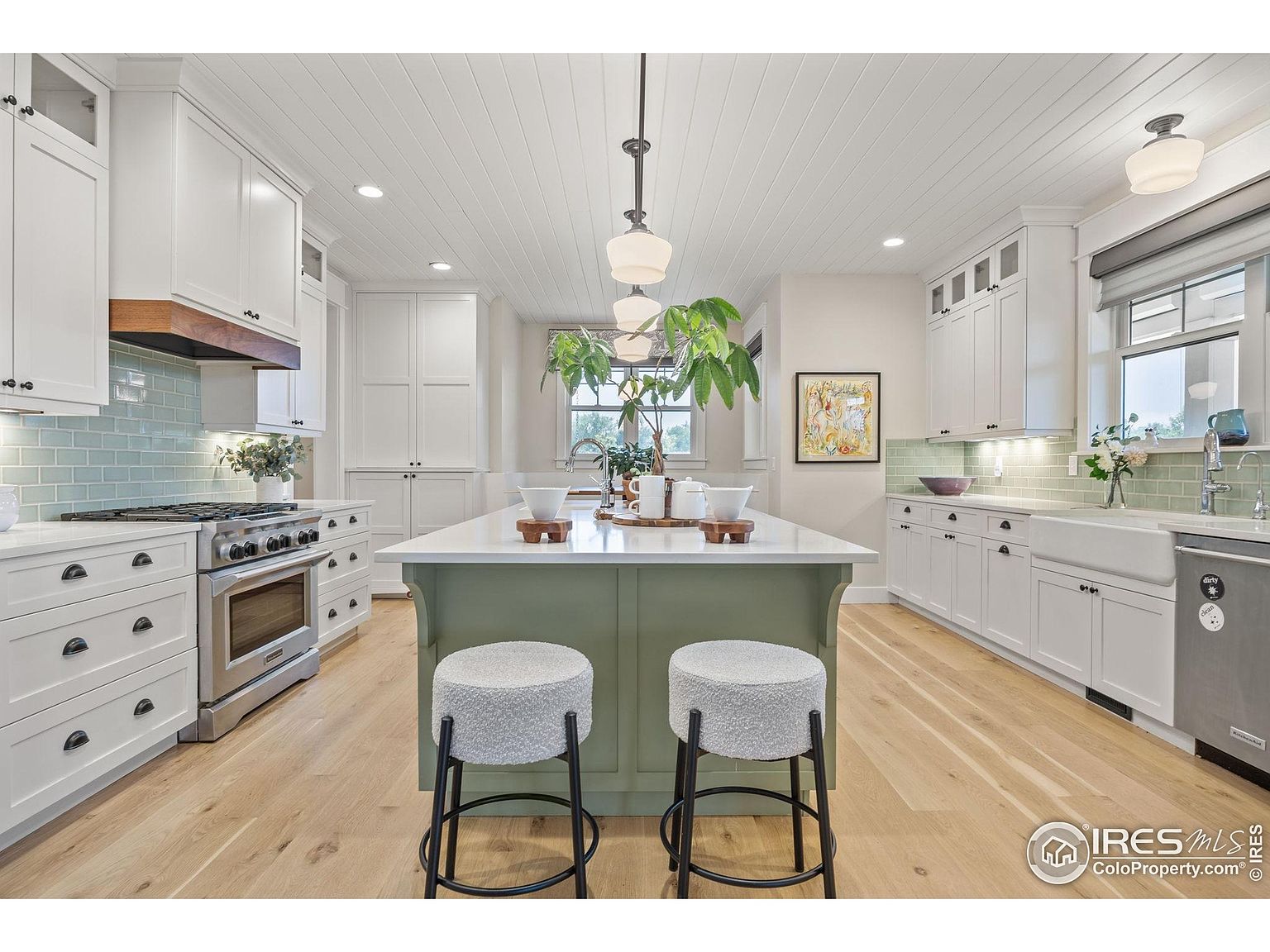This is a bright and airy kitchen featuring white cabinetry, stainless steel appliances, and a large green island with a white countertop. The kitchen has a farmhouse sink, light wood floors, and a subway tile backsplash. The perspective is a wide shot, showcasing the entire kitchen space.