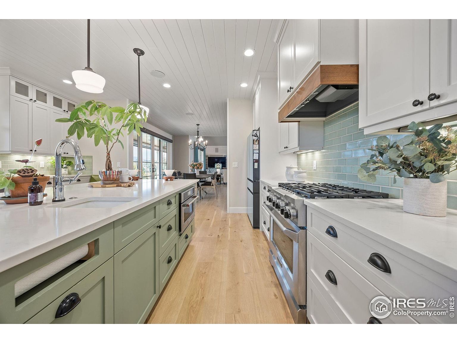 This is a well-lit kitchen featuring a large island with green cabinetry and a white countertop. Stainless steel appliances are visible, including a range and oven. The kitchen also has white upper cabinets and a light blue backsplash, creating a modern and inviting space.