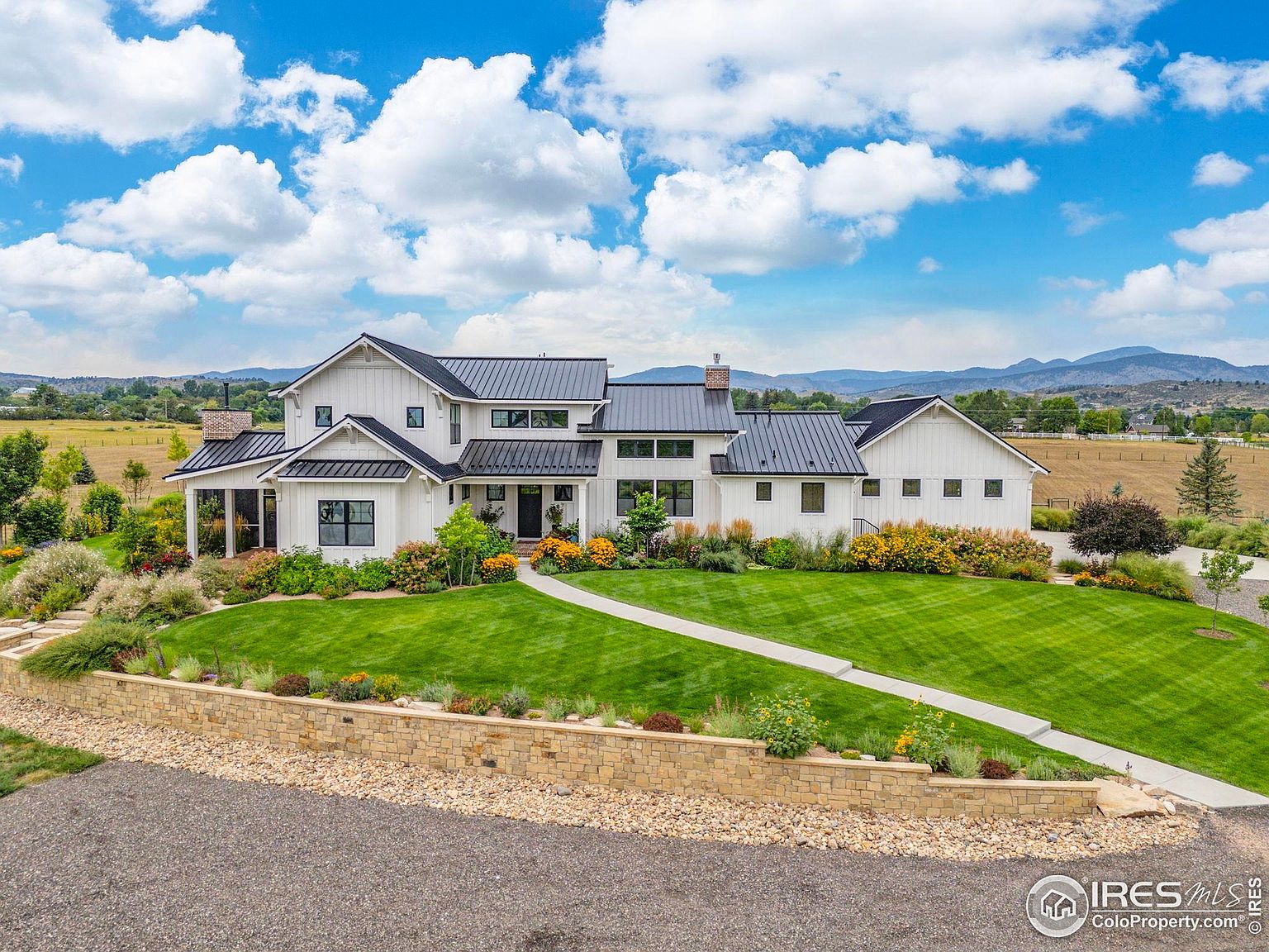 This is a stunning front view of a modern farmhouse-style home. The exterior features white siding, a dark metal roof, and black-framed windows. The professionally landscaped yard includes a stone retaining wall, lush green lawn, and colorful flower beds, creating a welcoming and picturesque setting.