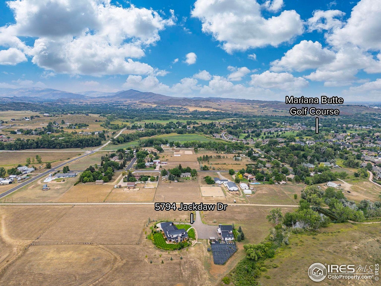 This aerial shot showcases a sprawling property featuring a modern home with dark roofing and light exterior, complemented by a smaller adjacent building. The landscape includes a well-maintained yard, surrounding fields, and distant mountain views under a partly cloudy sky. The image provides a comprehensive view of the property's setting and scale.