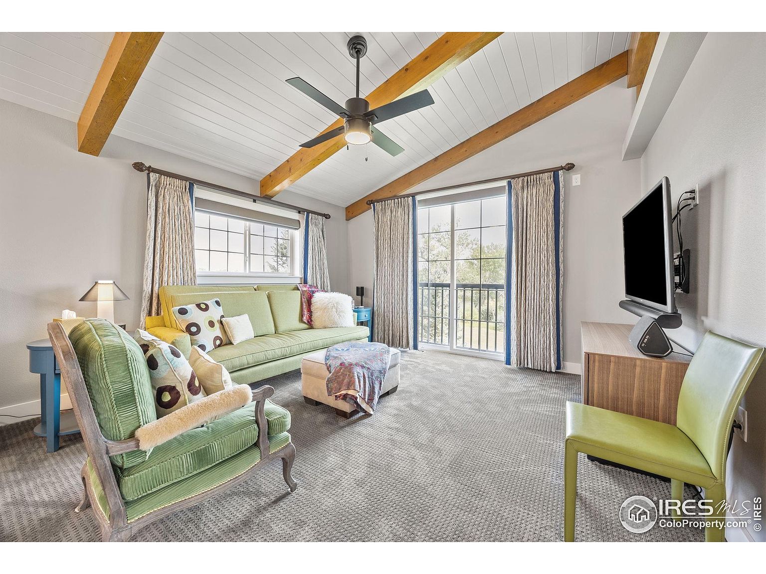 This is an interior shot of a living room featuring a vaulted ceiling with exposed wooden beams and a ceiling fan. The room is furnished with a green sofa, a green armchair, and an ottoman, creating a cozy and inviting atmosphere. Large windows and a sliding glass door provide ample natural light, enhancing the room's appeal.