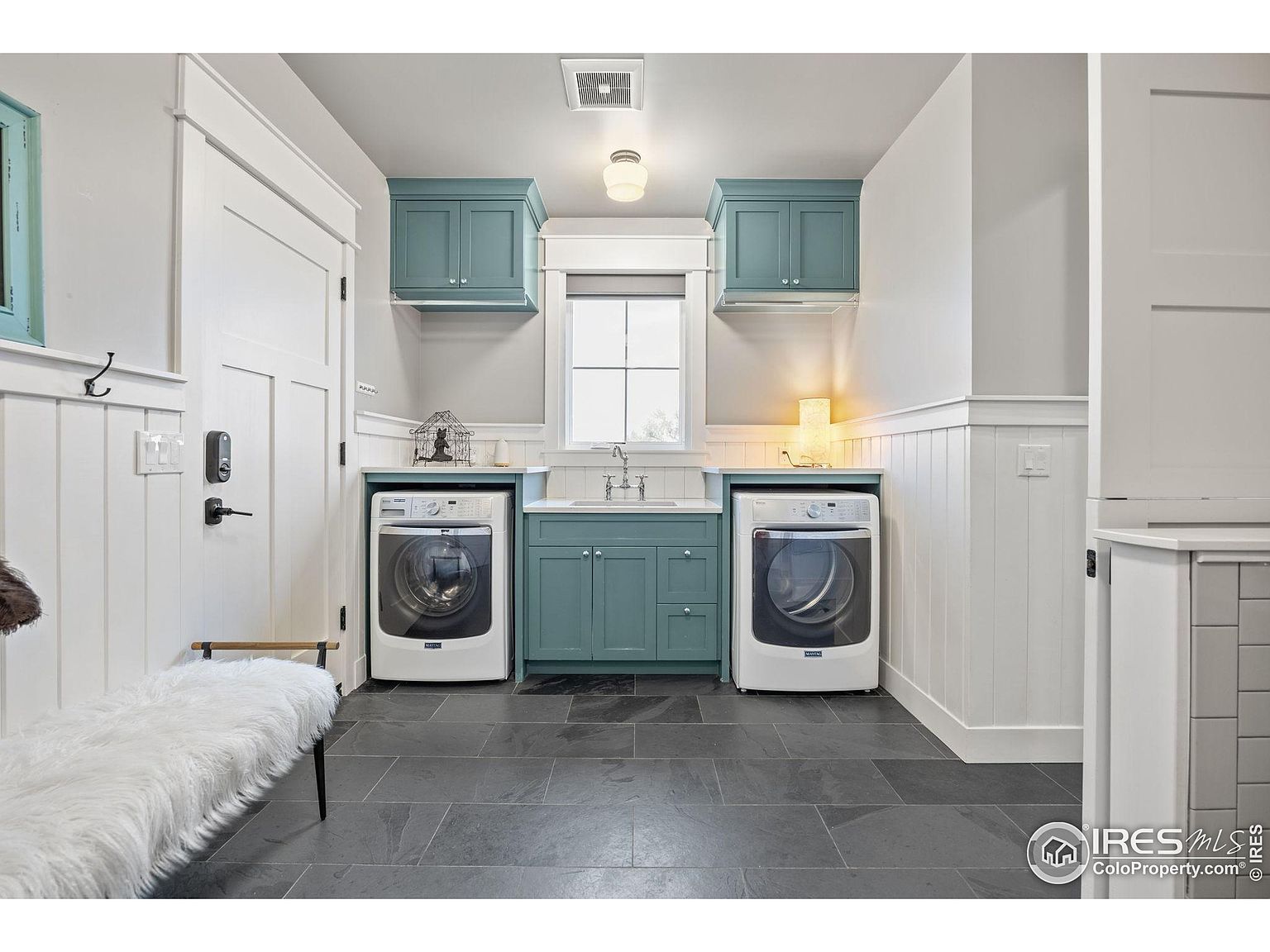 This is a well-organized laundry room featuring a modern design with teal cabinetry and white walls. The room includes a sink, countertop space, and front-loading washer and dryer units. The dark tile flooring adds a touch of sophistication, and the overall impression is clean and functional.