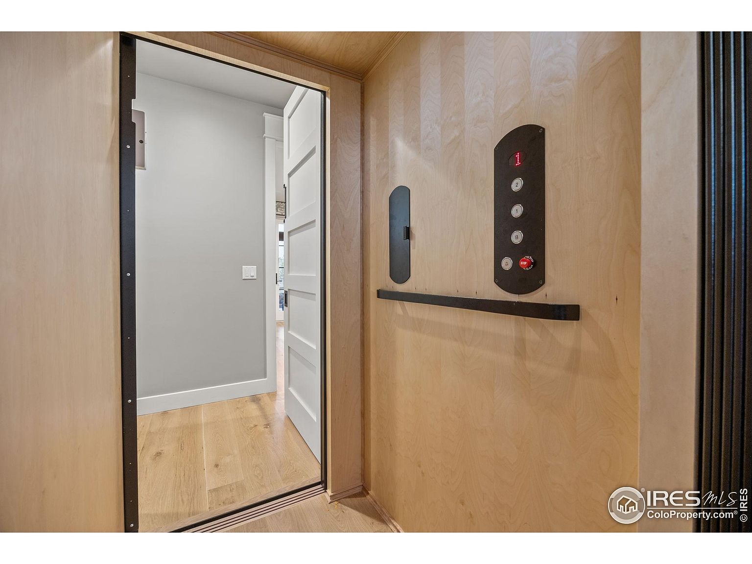 The image showcases an interior elevator, featuring light wood paneling and a control panel with buttons and a handrail. The elevator is positioned adjacent to a hallway with an open doorway, revealing a glimpse of a room with light gray walls and hardwood flooring. The overall impression is modern and functional, highlighting accessibility within the home.