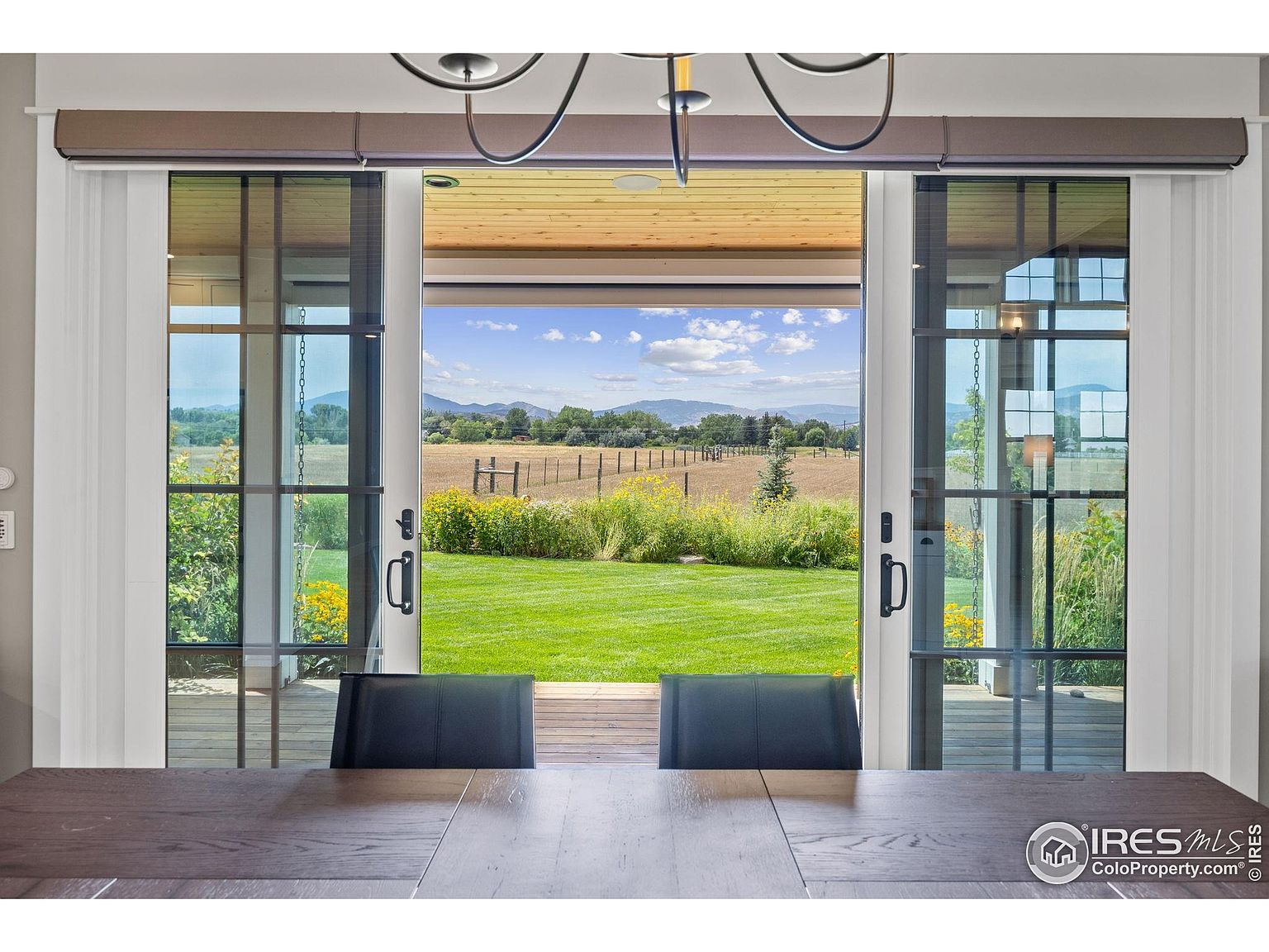 This image showcases a dining room with a view of a lush green yard and distant mountains through large sliding glass doors. The doors open onto a wooden deck, seamlessly blending indoor and outdoor living. A dark wood dining table and chairs are in the foreground, suggesting a comfortable and inviting space for meals and gatherings.