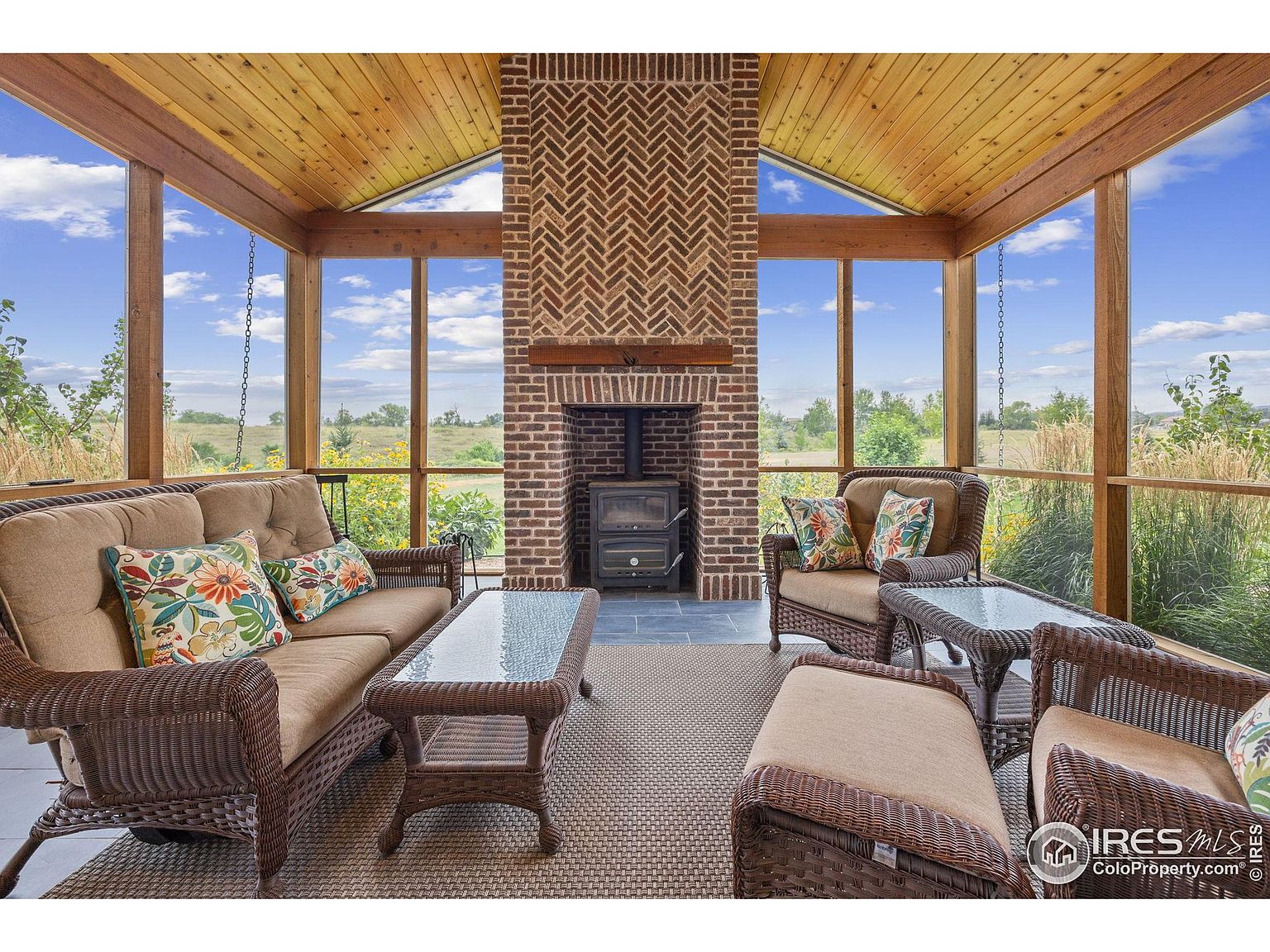 This sunroom features a brick fireplace as its focal point, complemented by wicker furniture including a sofa, chairs, and tables. Large windows offer expansive views of the surrounding landscape, creating a seamless connection with the outdoors. The room is bathed in natural light, enhancing the warm and inviting atmosphere.