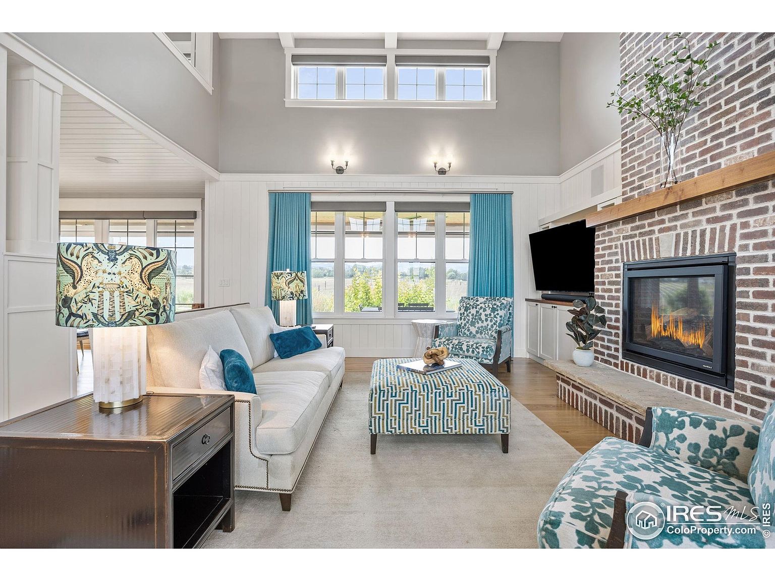This is a bright and airy living room featuring a brick fireplace with a wooden mantel and a flat-screen TV mounted above. The room is furnished with a light-colored sofa, patterned armchairs, and an ottoman, all arranged on a neutral-toned rug. Large windows with blue curtains provide ample natural light and a view of the outdoors, creating a comfortable and inviting atmosphere.