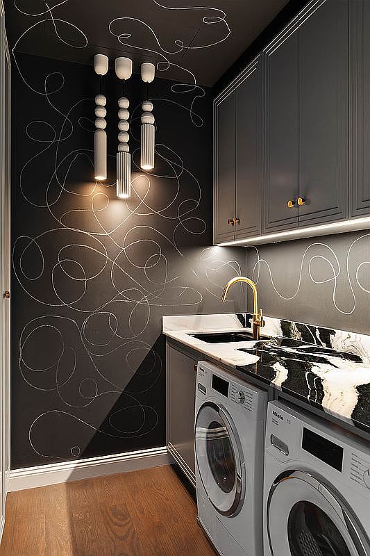 This is a modern laundry room featuring dark gray cabinetry with gold hardware and a black and white marble countertop. The walls are a dark color with a white abstract design, complemented by unique pendant lighting. Two front-loading washing machines are neatly installed under the counter, creating a functional and stylish space.