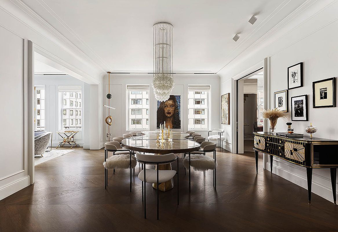 This is a spacious and elegant dining room featuring a large round table surrounded by stylish chairs with fur accents. A striking chandelier hangs above the table, and artwork adorns the walls, creating a sophisticated atmosphere. The dark wood flooring adds warmth and richness to the space, enhancing the overall luxurious feel.