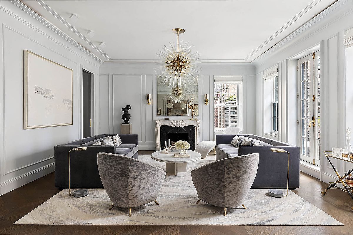 This is a beautifully staged living room featuring two gray sofas facing each other, complemented by two unique patterned armchairs. A round marble coffee table sits in the center of a light-colored area rug, beneath a striking starburst chandelier. The room is decorated with modern art and sculptures, and the walls are painted in a soft, neutral tone, creating an elegant and inviting atmosphere.