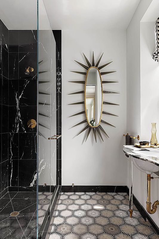 This is a stylish bathroom featuring a black and white color scheme. The shower is enclosed with glass and has black marble-like tiles with white veining, while the floor has a unique patterned tile. A starburst mirror hangs above a marble-topped sink with brass fixtures, adding a touch of elegance.