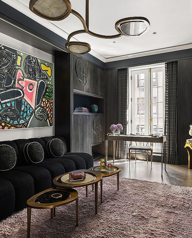 This interior shot showcases a sophisticated office or study with dark wood paneling and built-in shelving. A black sofa with round pillows sits adjacent to a set of nesting coffee tables, while a desk faces a window with sheer curtains. The room is illuminated by a modern chandelier, creating a luxurious and inviting workspace.