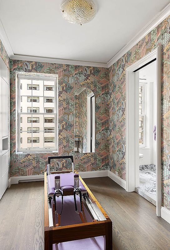 This interior shot showcases a home gym featuring patterned wallpaper and hardwood floors. A Pilates reformer is centrally positioned, and a window provides natural light. The room also includes a mirror and a doorway leading to another room, creating a functional and aesthetically pleasing workout space.