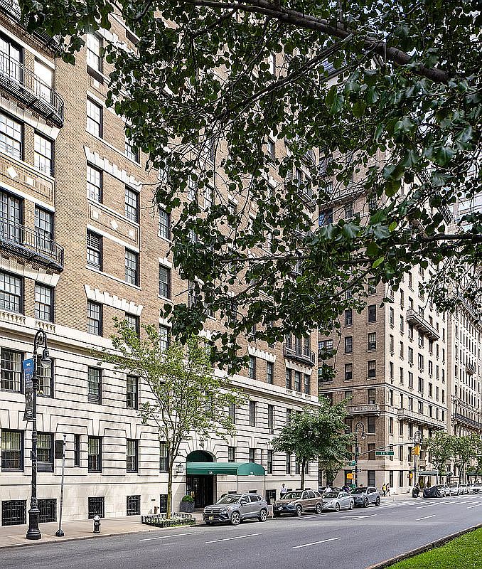 The image showcases the front exterior of a classic apartment building in an urban setting. The building features a mix of stone and brick facade, with symmetrical window placements and some balconies. A green awning covers the entryway, and the street in front is lined with parked cars and mature trees, creating a sophisticated and established atmosphere.