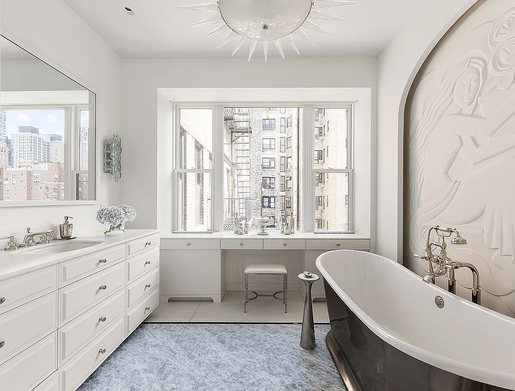 This is a luxurious primary bathroom featuring a freestanding bathtub with ornate fixtures, a vanity with ample storage, and a large window offering city views. An artistic wall relief adds a unique touch, while the blue-toned flooring complements the white cabinetry and walls. The overall impression is elegant and serene.